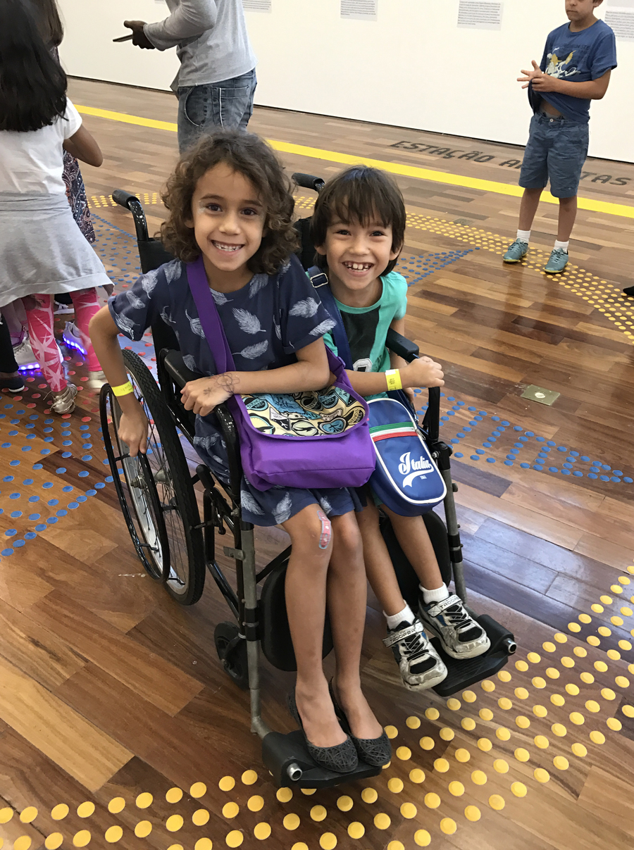 Tarsila e Lorenzo brincando com uma cadeira de rodas em uma exposição lúdica na Biblioteca Parque do centro do Rio | © Lima Andruška