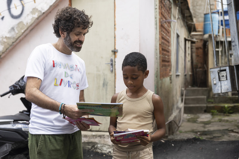 Vinicius Cortez, coordenador de marketing e comunicação da Bienal do Livro Rio, entrega livro para criança no Complexo da Alemão | © Bienal do Livro Rio